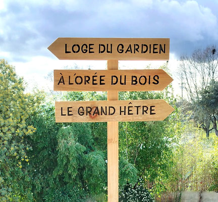 Totem fléché en bois recyclé brut, texte noir personnalisé pour guider les invités, planté dans un parc arboré