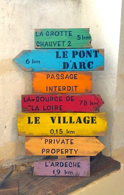 Panneau de direction multicolore, texte peint en noir personnalisé avec des lieux-dits et leur distance, posé sur un mur de pierre
