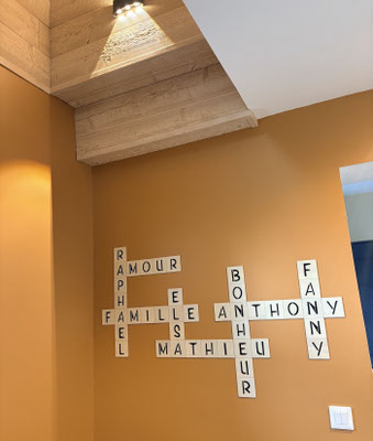 Lettres de Scrabble grand format collées sur un mur orange personnalisées avec des mots comme "famille" et "amour" et des prénoms