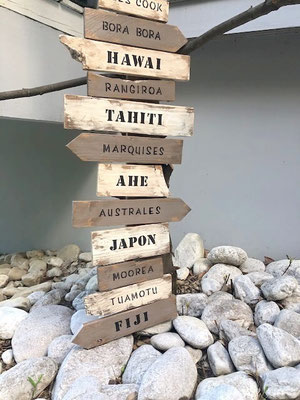 Panneau directionnel style bois flotté, avec plusieurs destinations comme "Hawaî", "Tahiti" ou "Japon" posé en extérieur sur des galets
