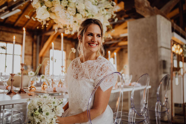 Braut sitzt lächelnd im Hochzeitsaal von Das Sägewerk, neben dekorierter Tafel mit Kerzen, weißen Blumen und transparenten Stühlen.