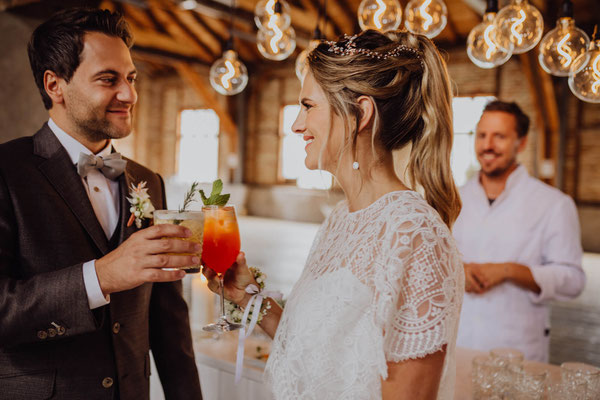 Brautpaar stößt mit Cocktails an der Bar im Hochzeitsaal von Das Sägewerk an, warme Glühbirnen-Lichter im Hintergrund.
