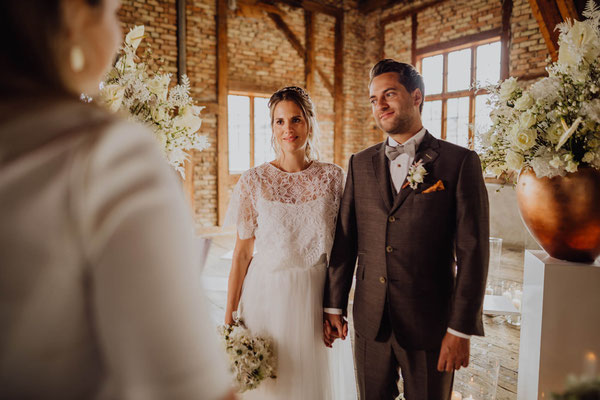 Brautpaar steht Hand in Hand während der Trauung im Hochzeitsaal von Das Sägewerk, umgeben von großen weißen Blumenarrangements.