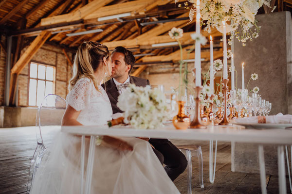 Brautpaar küsst sich am festlich gedeckten Hochzeitstisch im rustikalen Hochzeitsaal von Das Sägewerk mit Kerzen und Blumen.