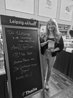 Malina Bura steht lächelnd und mit ihrem Buch in der Hand neben einem Aufsteller von Thalia in Leipzig.