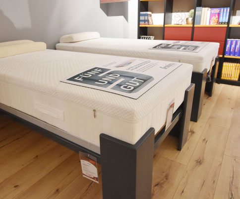Zwei graue Bettrahmen mit weißer Matratze auf einem Holzboden und Bücherregal im Hintergrund.