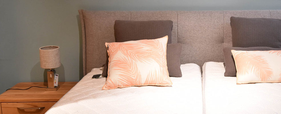 Boxspringbett mit grauem Stoffbezug, zwei Matratzen, weißem Topper und gemusterten Kissen.