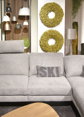 Graues Ecksofa mit Kopfstütze, beigem Kissen mit Aufschrift "SKI", zwei goldenen Wandkränzen.