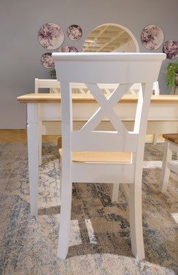 Nahaufnahme eines weißen Esszimmerstuhls mit gekreuzter Rückenlehne vor einem Holztisch.