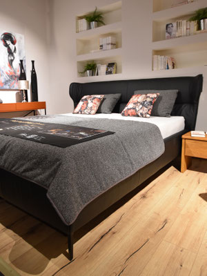 Ein Bett mit schwarzem Kopfteil, grauer Decke und gemusterten Kissen.