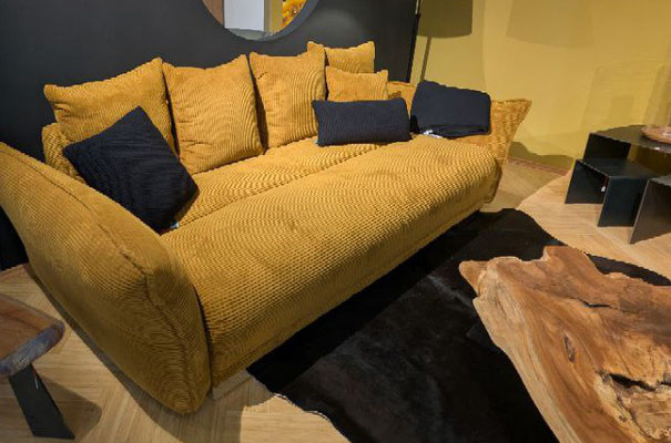 Senfgelbes Sofa mit dunklen Kissen neben rustikalem Holztisch auf dunklem Fellteppich.