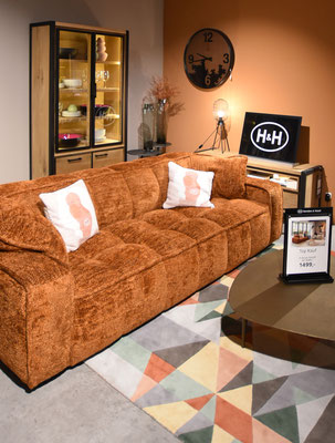 Braunes quadratisches Sofa mit zwei weißen Kissen und einer Vitrine im Hintergrund. Prozentzeichen.