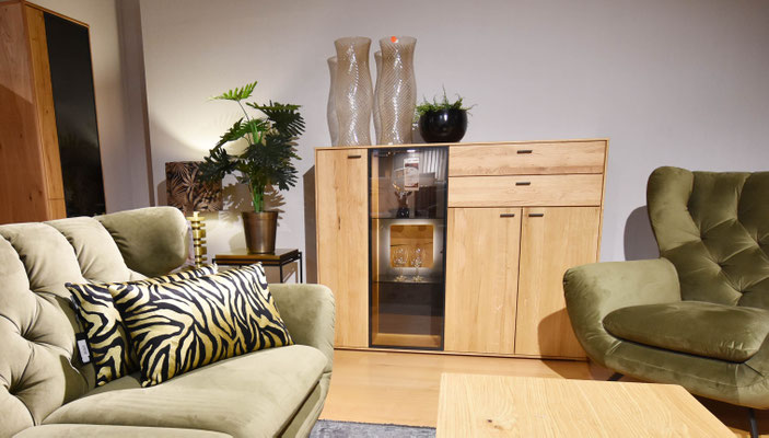 Links steht ein waldgrünes Sofa und rechts ein dazu passender Sessel. Dazwischen steht eine Holzkommode.