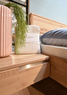 Schlafzimmer-Nachttisch aus Holz mit Beleuchtung, beigefarbener Bettwäsche und roséfarbener Vase.