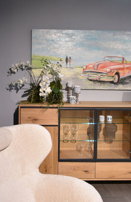 Modernes Sideboard aus hellem Holz mit Glasvitrine, einem Sessel, Orchideen und einem Gemälde.