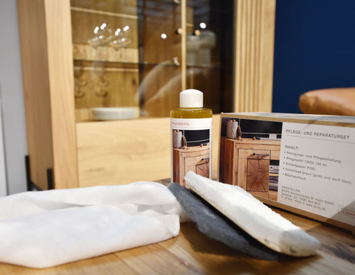 Pflegeset für Holzmöbel mit Ölflasche, Tuch, Schleifvlies und Schrank im Hintergrund.