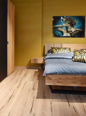Rechts steht ein Holzbett, die Wand dahinter ist senfgelb. Links steht ein Holzschrank.