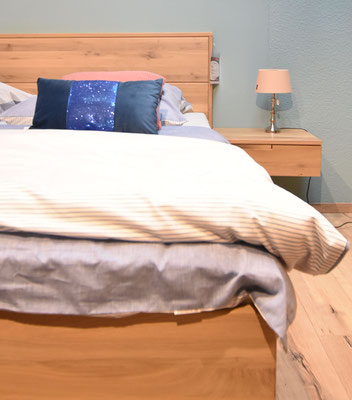 Ein Bett mit hellem Holzrahmen, gestreifter Bettwäsche und dunkelblauem Kissen mit Pailletten.
