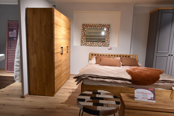 Modernes Schlafzimmer mit Holzschrank, Holzbett, heller Bettwäsche und Hocker mit Stoffbezug.