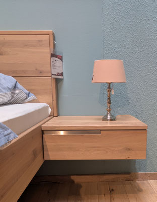 Neben einem Holzbett ein Nachttisch aus Eichenholz mit Schublade, darauf eine kleine Lampe.