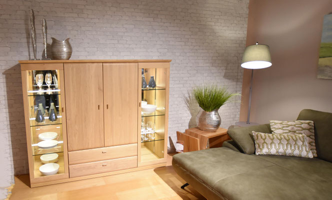 Helles Holzschranksystem mit Vitrine, daneben ein olivgrünes Sofa und eine Stehlampe.
