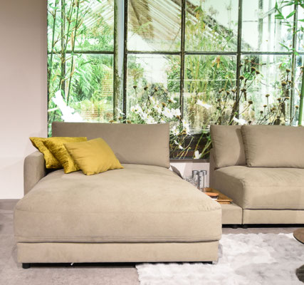 Beiges Chaiselongue-Sofa mit gelben Kissen vor einer Fototapete mit Gewächshaus-Optik.