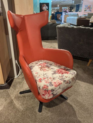 Orangeroter Sessel im Cockpit-Stil mit floral gemustertem Sitzpolster auf grauem Teppich.