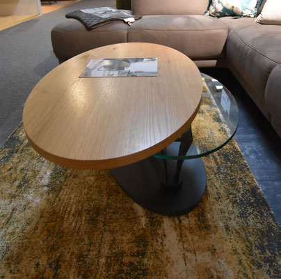 Runder Couchtisch mit Holzplatte und Glasplatte auf einem modernen Teppich vor einem Sofa.