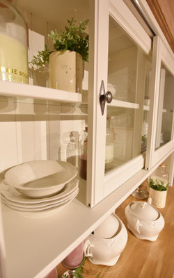 Weiße Glasvitrine mit Geschirr, Pflanzen und Kerzen im Landhaus-Stil.