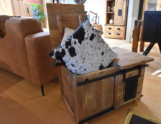 Hölzerne Truhenbank mit schwarz-weißem Kuhfellkissen neben einem braunen Sofa.