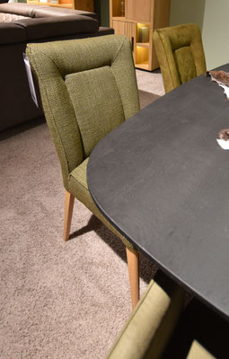 Olivgrüner Stuhl mit Stoffbezug und hellen Holzbeinen, neben dunklem Tisch mit schwarzer Oberfläche.