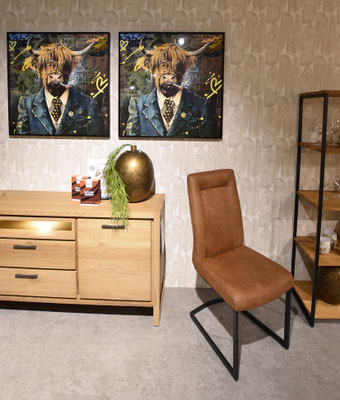 Brauner Freischwinger-Stuhl mit schwarzem Metallfuß vor einem Sideboard und Pop-Art-Bildern.