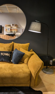 Ein senfgelbes Sofa mit Kissen. Dahinter ist ein runder Spiegel und eine Stehlampe zu sehen.