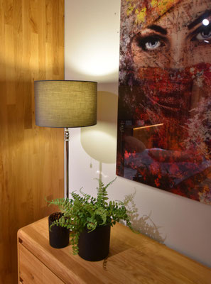 Silberne Tischlampe mit grauem Schirm und Farn in schwarzem Topf auf einer Kommode neben Kunst.