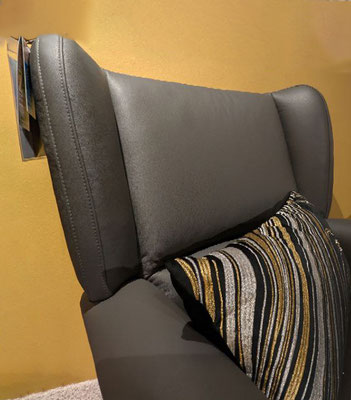 Rückenlehne eines grauen (Leder-)Sessels mit Ohrenbacken und einem schwarz-gold gestreiften Kissen.