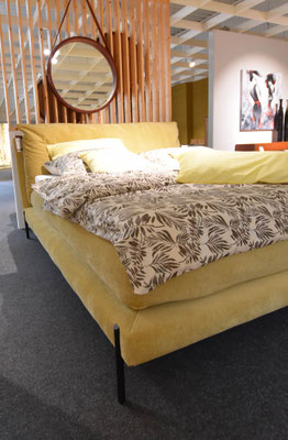 Senffarbenes Boxspringbett auf schwarzen Füßen mit gemusterter Bettwäsche vor Holzwand und Spiegel.