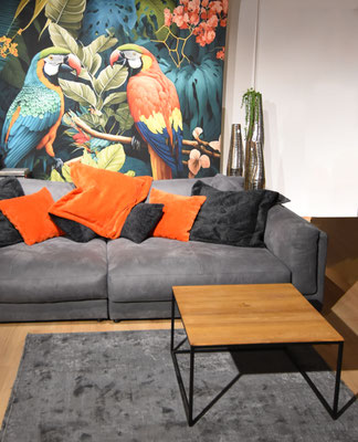 Graues Sofa mit bunten Kissen, Holztisch, grauer Teppich und eine tropische Tapete mit Papageien.