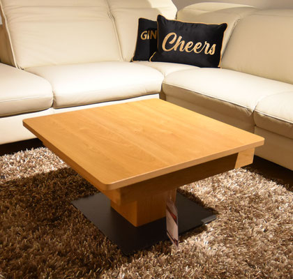 Quadratischer Holztisch mit schwarzem Fuß auf braunem Teppich vor einem cremefarbenen Sofa.