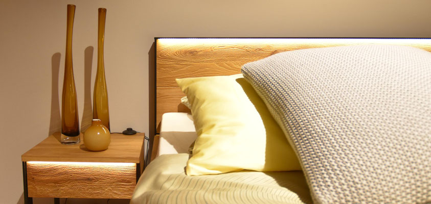 Beleuchteter Holznachtschrank mit drei Vasen neben einem Holzbett mit Kissen.