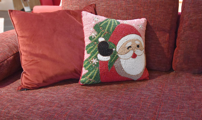 Stickkissen mit Weihnachtsmann-Motiv und Tannenzweig auf roter Couch und weitere rote Kissen.