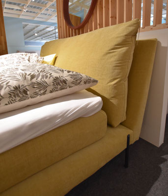 Blick auf ein großes, gelbes Boxspringbett mit vielen Kissen und floral gemusterter Bettwäsche.
