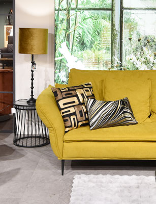 Senfgelbes Sofa mit Musternkissen, goldener Samt-Tischlampe und einem Bild einer grünen Szenerie.