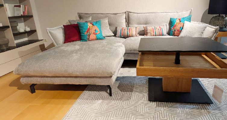 Graues Ecksofa mit bunten Kissen neben einem modernen Holztisch in einem hellen Wohnzimmer.