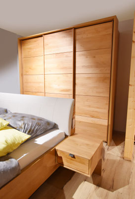Nahansicht eines Holzbettes mit grau-gelber Bettwäsche und weißem Kopfteil. Nachttisch und Schrank.
