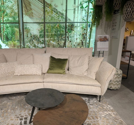 Beiges Sofa mit Kissen, einem runden Couchtisch-Set und einer großen Fensterfront mit Pflanzen.