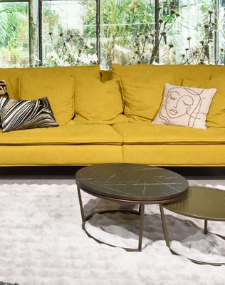 Gelbes Sofa mit Kissen, ovaler Couchtisch, hellgrauer Teppich vor Fenster mit Blick ins Grüne.