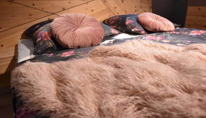 Holzbett mit dunkler Blumen-Bettwäsche, zwei runden Kissen in Altrosa und einer flauschigen Decke.