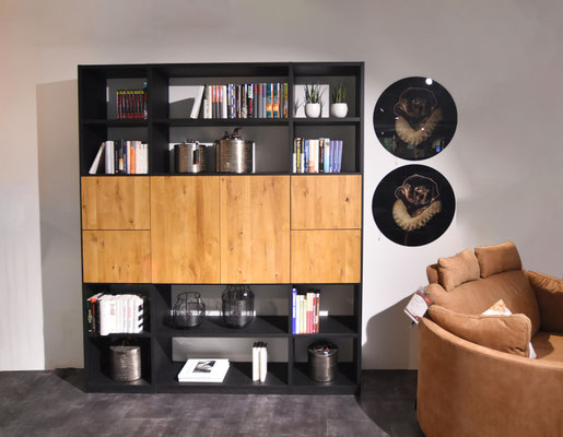 Ein dunkelbraunes, rundes Sofa mit braunen Kissen steht vor einem schwarz-holzfarbenen Regal.