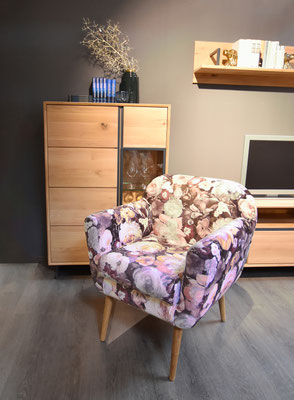 Ein Sessel mit buntem Blumenmuster steht vor einem hellbraunen Holzschrank.