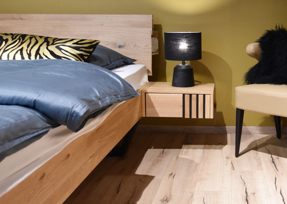 Ausschnitt eines Schlafzimmers mit Holzbett und schwarzer Nachttischlampe, schlicht modern gestaltet.